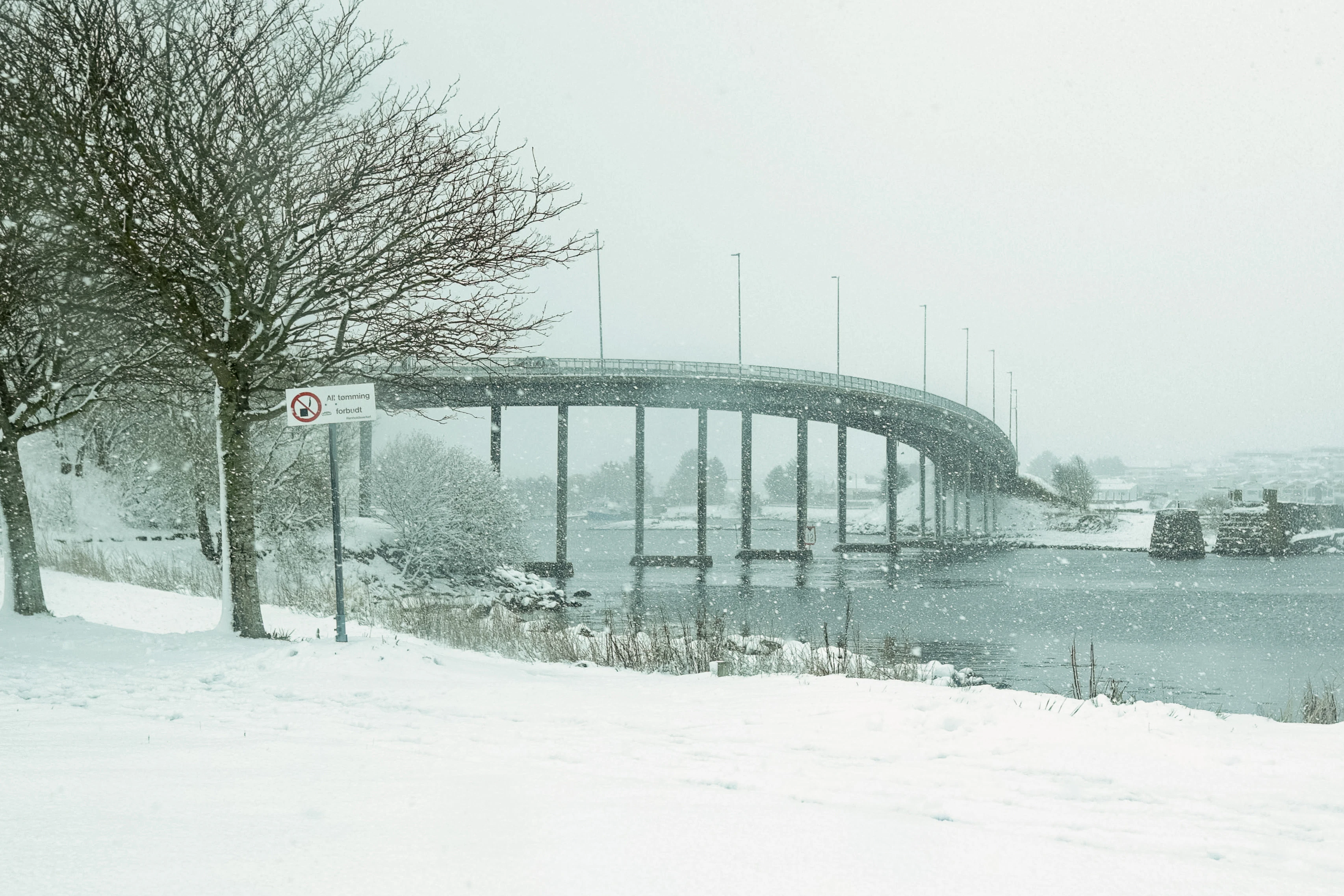 Snow falls on a bridge over water.